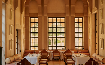 Amanbagh restaurant interior with warm stone walls, tall arched windows, and wooden dining chairs arranged throughout the space.