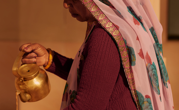 A woman at Amanbagh holds a brass watering can, wearing traditional Indian attire in deep red with a patterned shawl.
