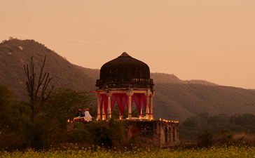 Chhatri dinner pavilion at Amanbagh, illuminated at dusk in a mustard field with mountains beyond.