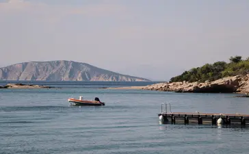 Wooden pier extending into clear turquoise waters at Amanzoe, Greece, with a small boat and distant cliffside visible.