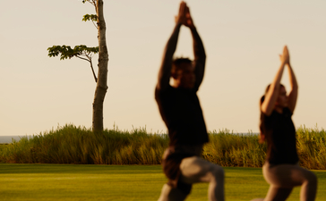 Deux personnes pratiquent le yoga en plein air à Amanera, la station balnéaire, avec des arbres et un terrain verdoyant en arrière-plan.
