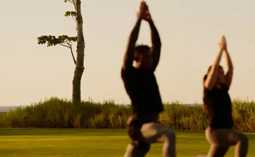 Duas mulheres a fazer yoga ao ar livre no resort Amanera, com paisagem verde ao fundo.