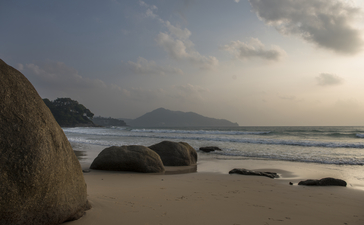 Granite boulders scattered across a sandy beach at Amanpuri, with calm waters and distant headlands under soft evening light.