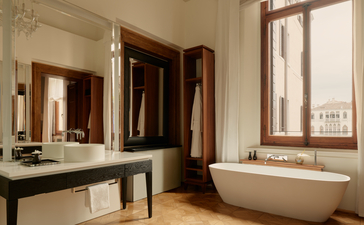 アマン ヴェニスのバスルーム。大理石のカウンターと鏡、キャナル・グランデの景色を眺める窓、白い浴槽。