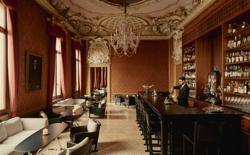Bar at Aman Venice with warm terracotta walls, ornate ceiling, and shelves of spirits behind the counter.
