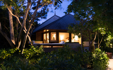 Amanpulo villa bedroom illuminated at dusk, nestled amongst tropical foliage and towering trees.