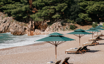 Sunlit beach at Aman Sveti Stefan with loungers beneath parasol, turquoise waters and rocky cliffs beyond.