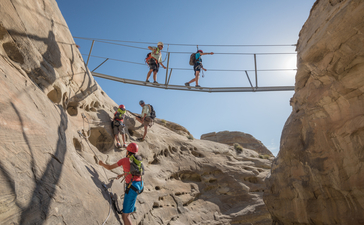 Family rock climbing on via ferrata at Amangiri, Utah, with climbers navigating a suspended bridge between sandstone formations.