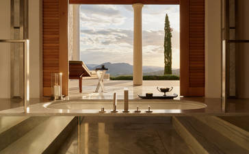 Bathroom at Amanzoe with limestone surfaces, candles, and sea views through large windows.