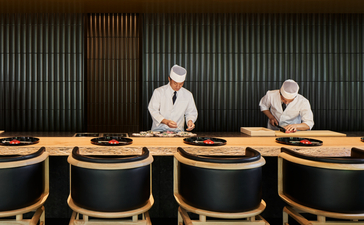 Two chefs stand behind a wooden counter at Aman Kyoto, with empty dining chairs lined in front.