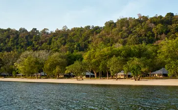 Amanwana's beachfront with sandy shore, tropical forest canopy, and thatched structures along the waterline.