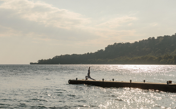 Wooden pier extending into calm waters at Amanwana, Indonesia, with forested coastline beyond.