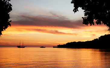Sunset over the sea at Amanwana, with a sailing vessel on the water and forested shoreline silhouetted against golden sky.
