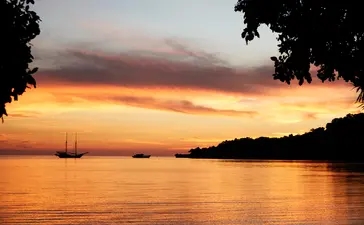 Sunset over the sea at Amanwana, with a sailing vessel on the water and forested shoreline silhouetted against golden sky.