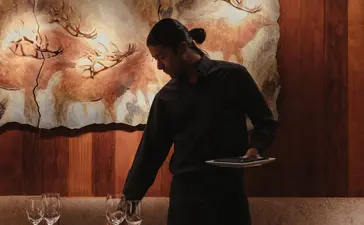 Server in black uniform arranging table settings in Amangani's dining venue, warm wood interior visible behind.