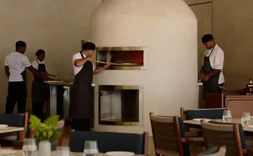Amanera resort's beach club interior with guests gathered around a central fireplace, wooden dining furniture, and natural light from windows.