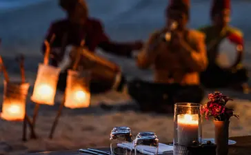 Candlelit dinner table at Amanwana with guests gathered on the beach at dusk in Sumbawa, Indonesia.