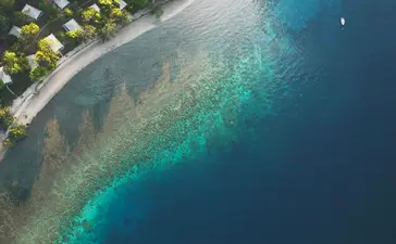 Aerial view of Amanwana's sandy beach curving along turquoise waters, with thatched bungalows nestled among tropical vegetation.