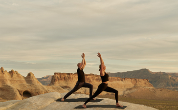 Two people practising yoga on a rock formation at Amangiri, with desert landscape and mountains in the background.