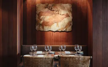 Amangani dining venue with woven chairs facing a warm-lit stone fireplace.