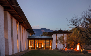 Amankora Bhutan lodge exterior at dusk, with warm interior lighting glowing through large windows and mountains beyond.