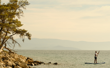 Person paddleboarding in calm turquoise waters at Amanruya, Turkey, with rocky shoreline and tree in foreground.