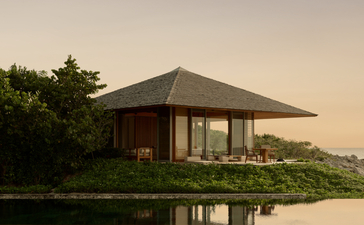 The Artist's Villa at Amanyara, a timber-clad pavilion with pitched roof, reflected in still water at dusk.