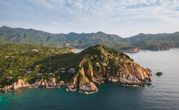Amanoi's dramatic rocky headland rises from turquoise waters, framed by forested mountains and misty horizon.