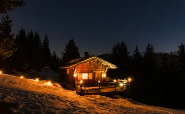 Wooden chalet glowing warmly at dusk surrounded by snow and evergreen trees at Aman Le Mélézin resort.