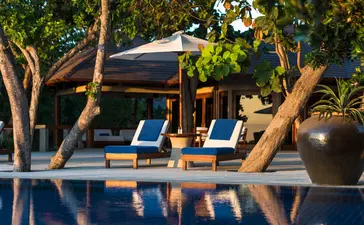 Wooden lounge chairs beside an infinity pool at Amanpulo resort, framed by tropical trees at golden hour.