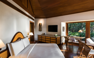 Bedroom at Amanpulo resort with wooden ceiling, white walls, bed with yellow accents, and views through glass doors to tropical surroundings.