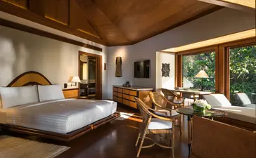 Chambre spacieuse au resort Amanpulo avec lit king-size, mobilier en bois naturel et grandes fenêtres donnant sur la végétation.