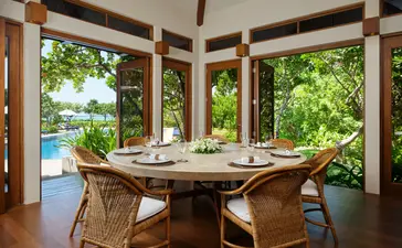 Dining room at Amanpulo resort with wooden table, rattan chairs and open views to tropical garden and water beyond.