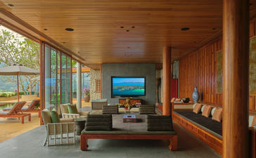 Amanpuri accommodation living room with warm wood panelling, floor-to-ceiling windows overlooking tropical landscape, and modern furnishings.