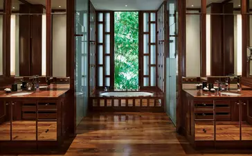Symmetrical bathroom with dual wooden vanities, central glass doors opening to greenery, and warm toned cabinetry at Amanpuri.