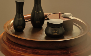 Aman Essentials skincare products displayed on a wooden round table at Amantaka, featuring black bottles and a cream jar.