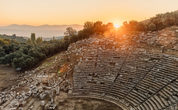 Ancient Greek theatre at Amanruya at sunset, with stone seating rows descending towards the stage, bathed in golden light.