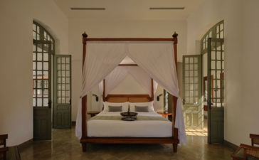 Amantaka principal bedroom with four-poster bed, symmetrical windows, and wooden shutters opening to Mekong views.