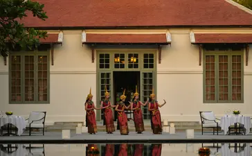 Monks in saffron robes stand at the entrance of a colonial building at Amantaka, with poolside dining setup in the foreground.
