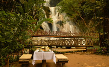 Outdoor breakfast table at Amantaka with Kuang Si Waterfall backdrop and lush greenery.
