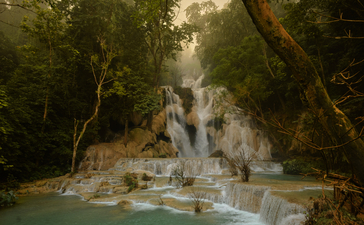 Tiered limestone waterfall flowing through lush forest at Kuang Si, Laos.