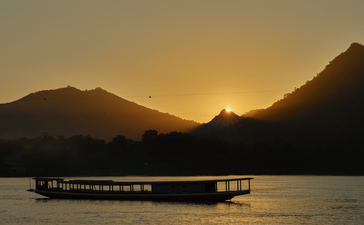 Wooden boat on the Mekong River at sunset, with mountains silhouetted against golden sky at Amantaka.