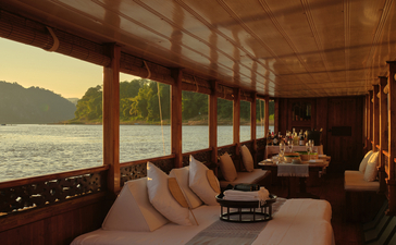 Amantaka dining space with floor-to-ceiling windows overlooking the Mekong River at sunset, featuring wooden furnishings and warm lighting.