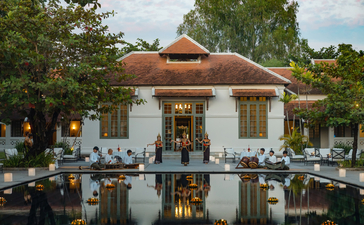 Amantaka's ochre-coloured exterior reflected in still water, framed by tropical trees.