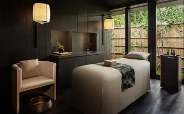 Salle de soins spa apaisante à Aman Kyoto avec lit de traitement, fauteuil crème et grandes baies vitrées donnant sur un jardin.