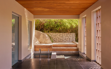 Amanoi resort lake wellness pool villa interior, viewed through open doors towards a wooden bench on a terrace overlooking water and stone wall.