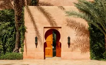 Architectural detail at Amanjena resort showing a terracotta archway with golden doors reflected in still water, flanked by palm trees.