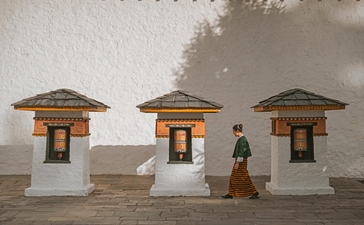 Amankora's Thimphu Lodge features three traditional Bhutanese stone cottages with dark roofs and wooden doors, a figure standing between them.