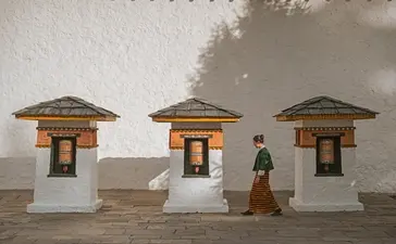 Amankora's Thimphu Lodge features three traditional Bhutanese stone cottages with dark roofs and wooden doors, a figure standing between them.