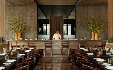 Dining room at Aman New York with two tables, dark wood chairs, and a central bar with shelving and golden branches in tall vases.